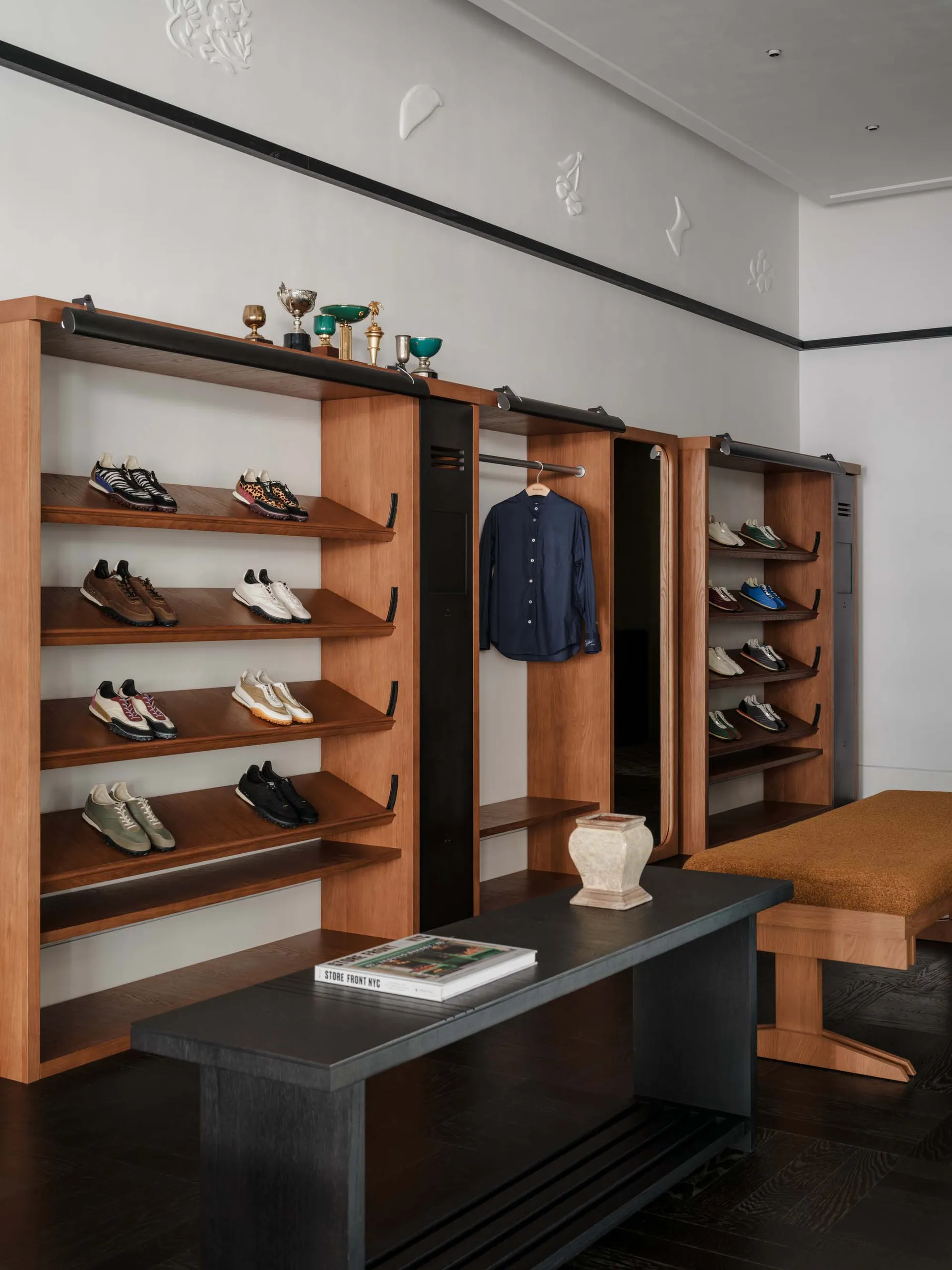 Modern walk-in closet with walnut open shelving displaying numerous sneakers, a navy shirt hanging, dark console table, and upholstered bench in a luxurious dressing room.