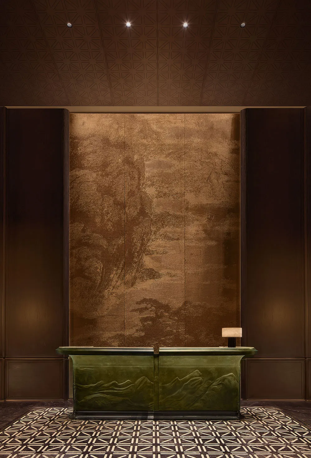 Luxurious hotel or corporate reception desk featuring a carved malachite-green front with mountain relief, set against a large bronze-toned Chinese landscape mural, under a geometric coffered ceiling with patterned marble floor.