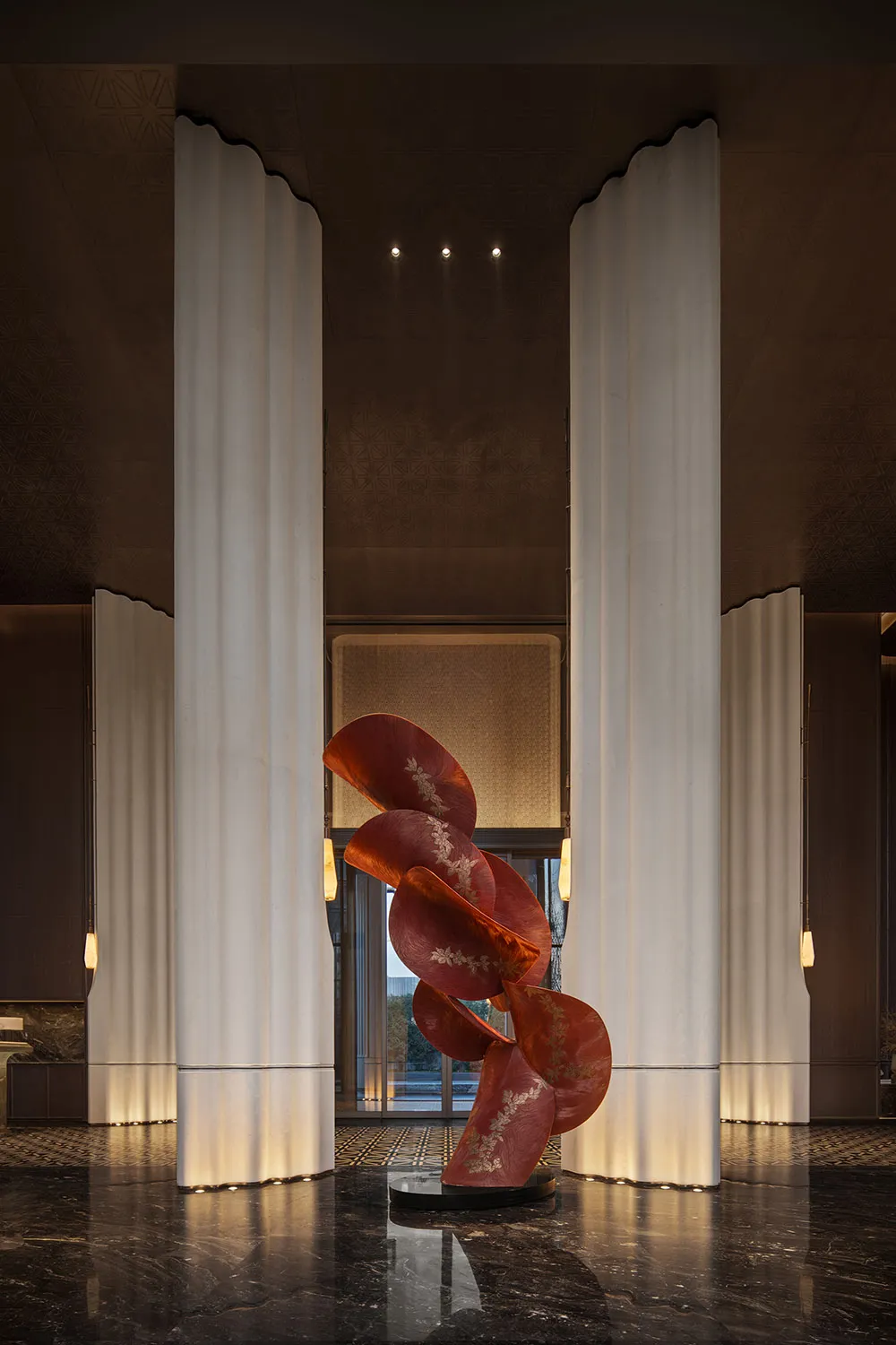 Luxurious hotel lobby featuring a large abstract red sculpture with gold detailing, flanked by tall white fluted marble columns on a polished dark marble floor.