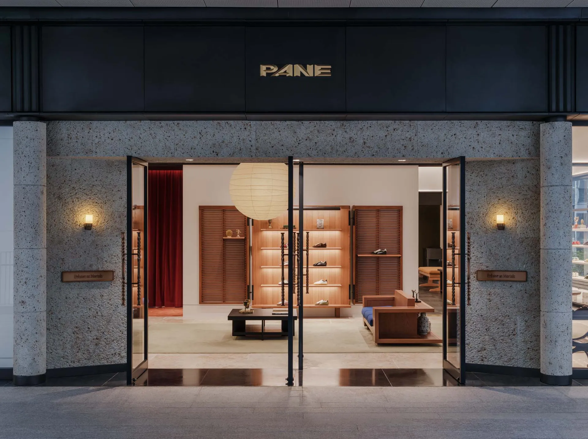 Front entrance view of PANE shoe store featuring large terrazzo pillars, black metal-framed glass doors, wooden display shelves with integrated lighting, and a large white paper lantern pendant.