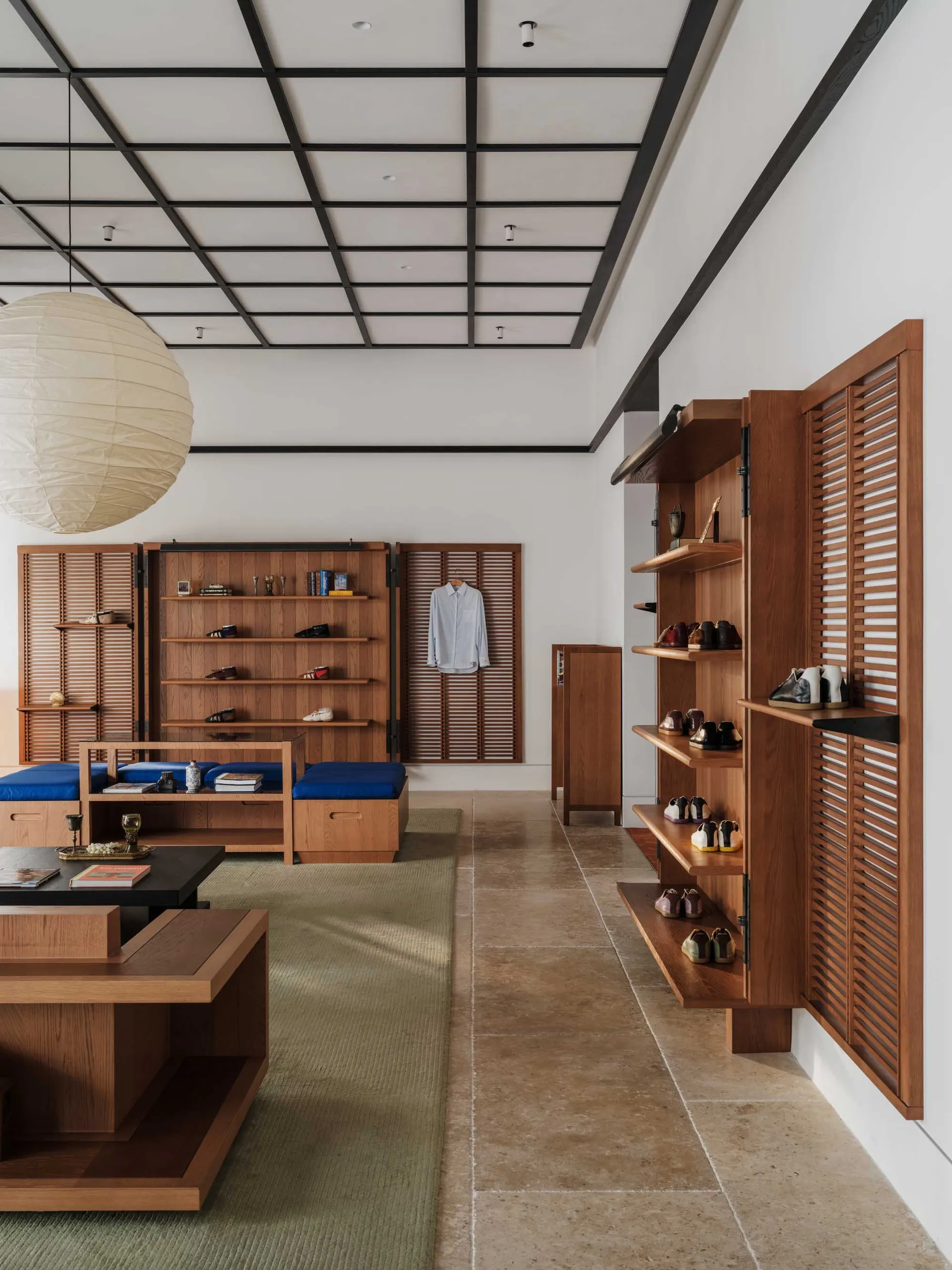 Modern Japanese-style entryway featuring warm teak wood open shelving for shoes, built-in blue cushioned seating, a large paper lantern pendant, and tatami-style green rugs on a stone tile floor.