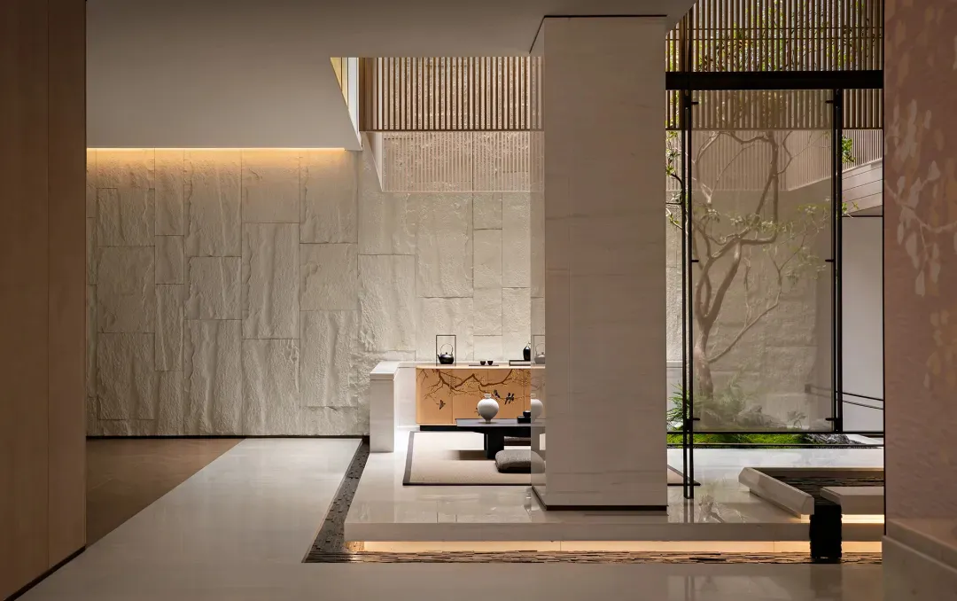Modern Japanese minimalist tea room featuring a large textured limestone wall, warm cove lighting, wooden console with tea set, and direct view to a lush green courtyard garden through floor-to-ceiling glass.