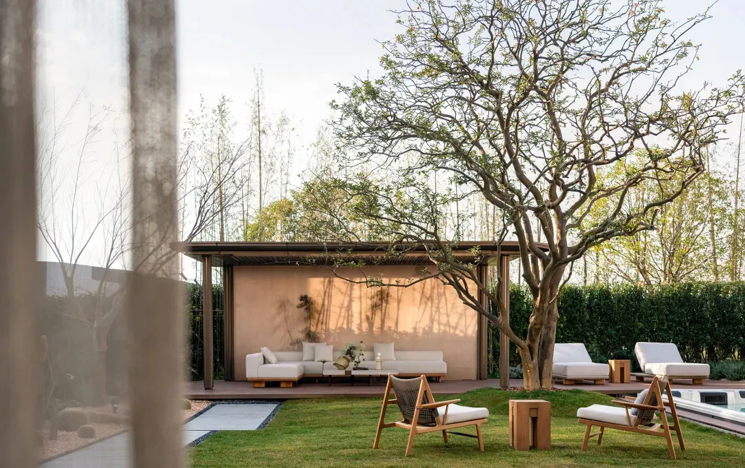 Modern outdoor living space featuring wooden lounge chairs, white sectional sofa, peach-colored wall, and a large deciduous tree on a manicured lawn.