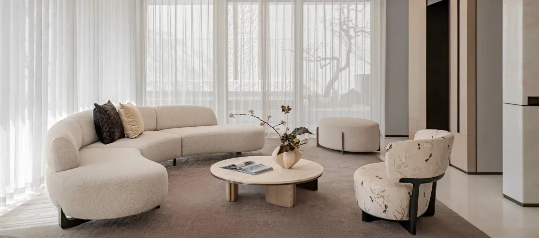 Modern minimalist living room featuring a large cream boucle curved sectional sofa, matching floral accent armchair, round light-wood coffee table, and sheer white curtains allowing abundant natural light.
