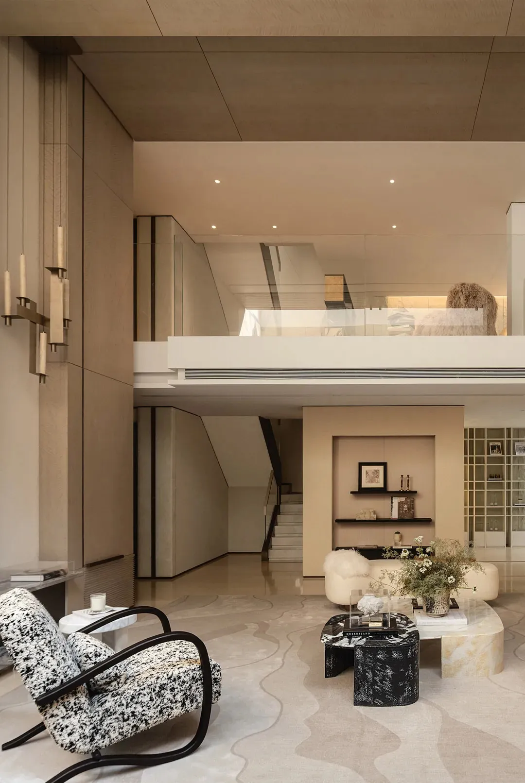 Modern double-height living room with a black and white patterned armchair, marble and terrazzo coffee tables, cream curved sofa, and a second-floor mezzanine with glass railing.