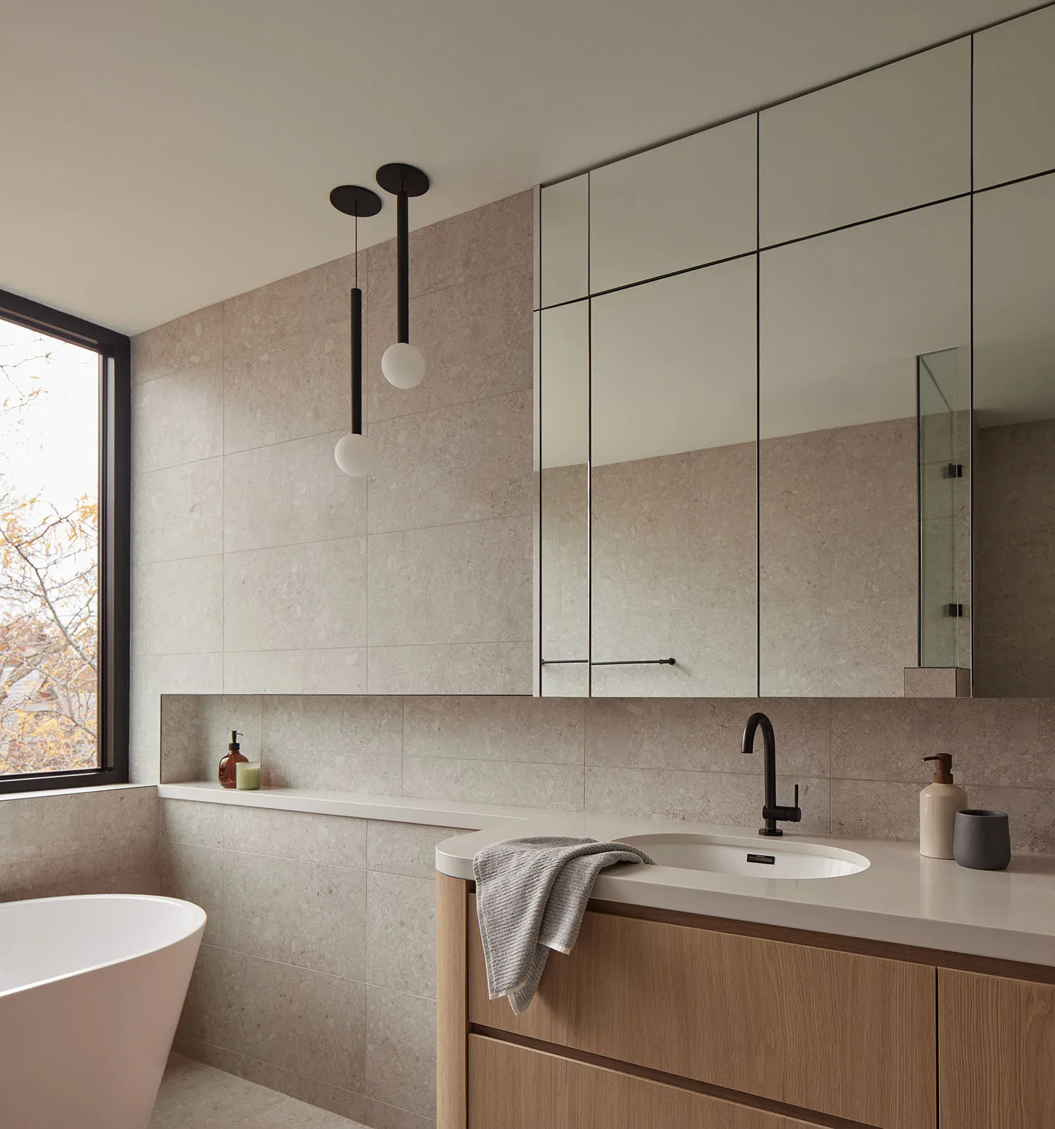 Modern minimalist bathroom with large-format travertine tile walls, floating light oak vanity with white countertop, black matte faucet, integrated mirror cabinet, and two black pendant lights with white glass globes above a freestanding white bathtub.