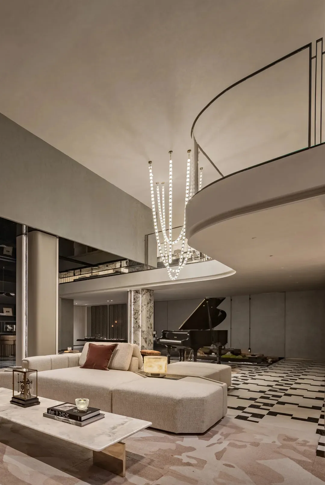 Modern luxury double-height living room with beige sectional sofa, marble coffee table, black grand piano, and cascading crystal chandelier under a curved mezzanine.
