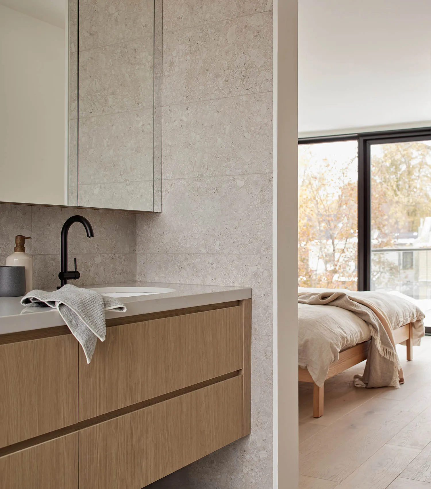 Modern bathroom featuring a travertine tiled wall and countertop, light oak floating vanity with black matte faucet, and a view into a minimalist bedroom with wooden bed and large window.