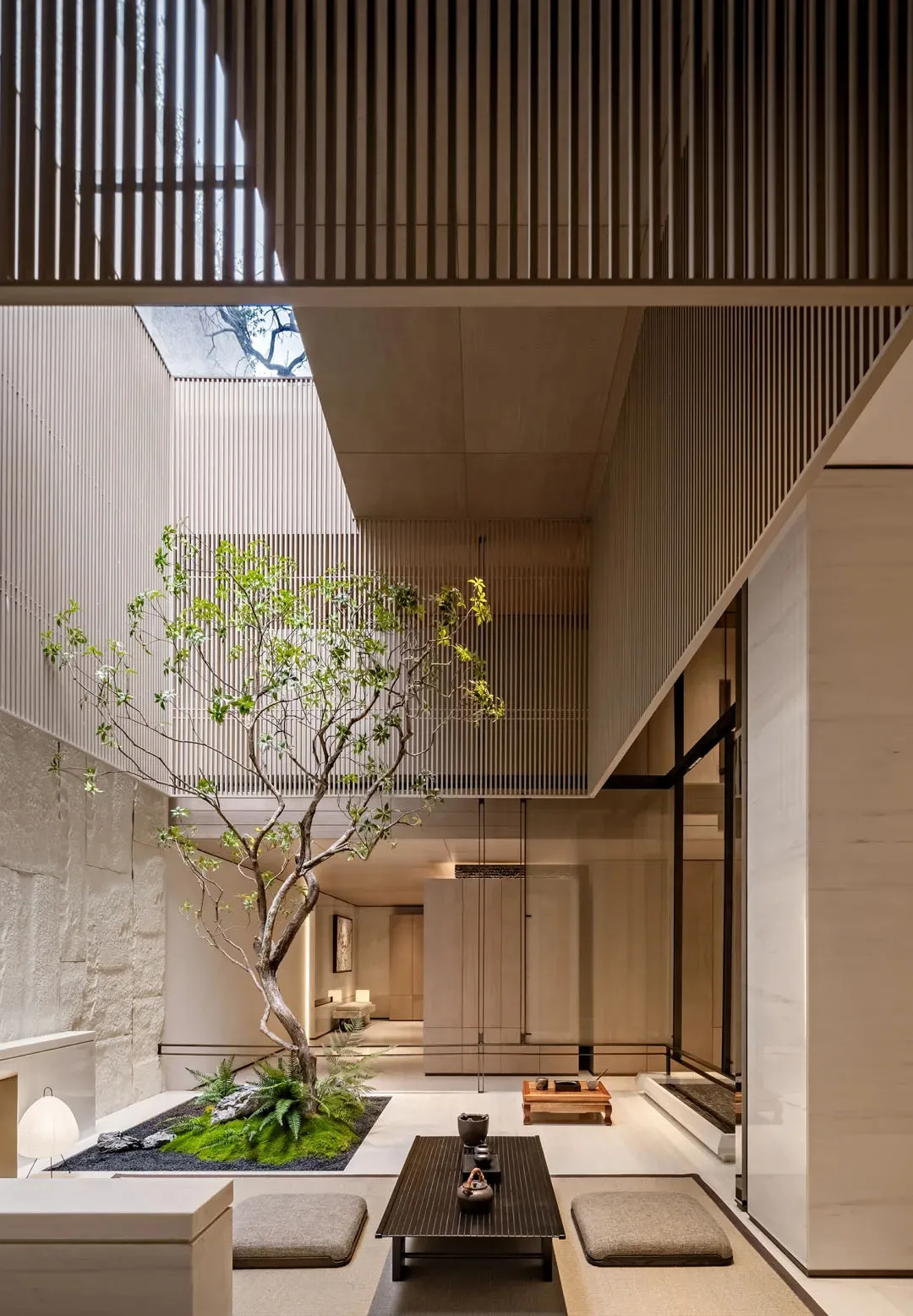 Modern Japanese minimalist indoor courtyard featuring a living tree, moss garden, skylight, wooden tea table, and floor cushions within a concrete and wood architectural space.