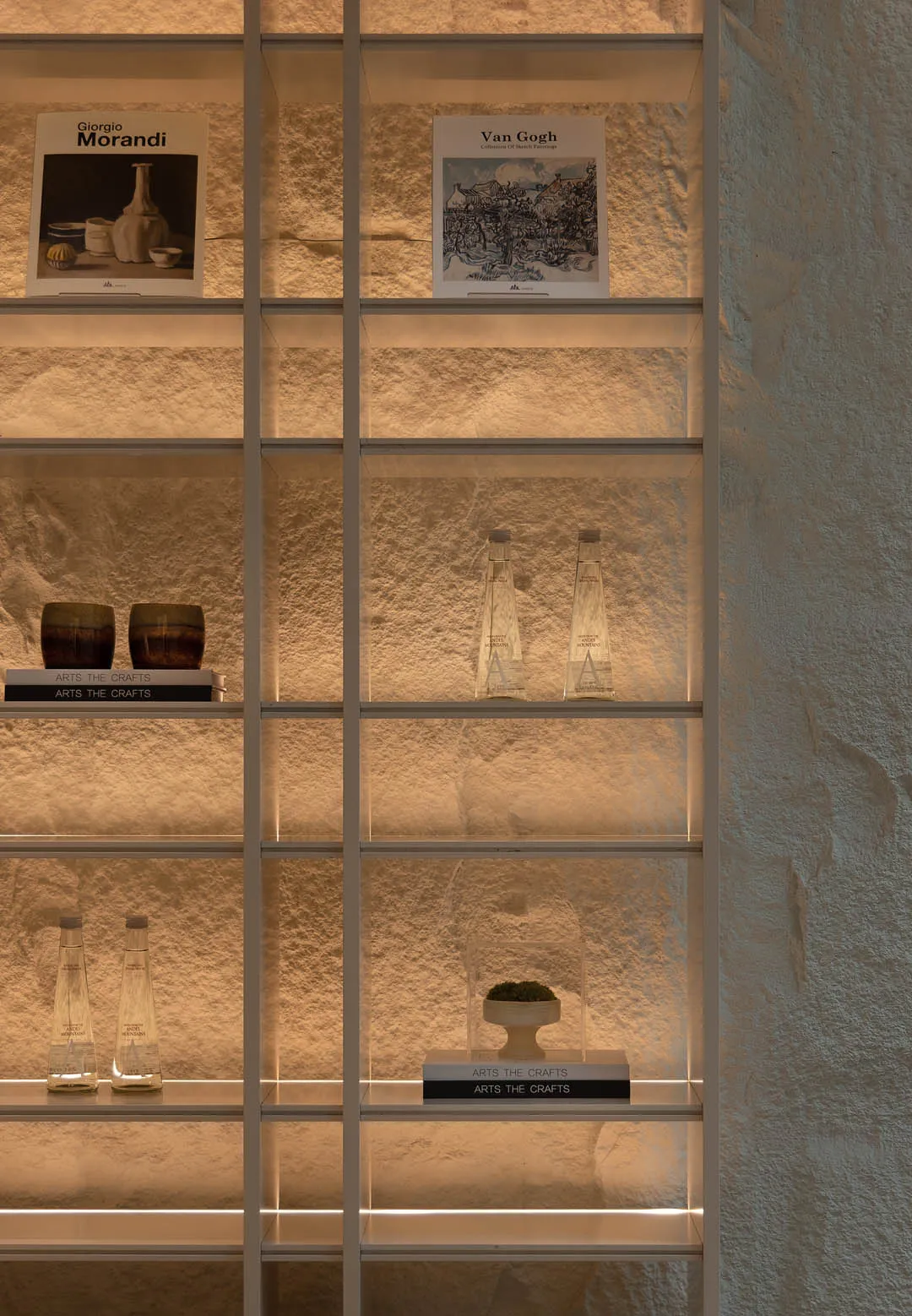 Built-in white shelving unit with warm backlighting showcasing art books, ceramic cups, glass bottles, and a moss sculpture against a textured plaster wall.