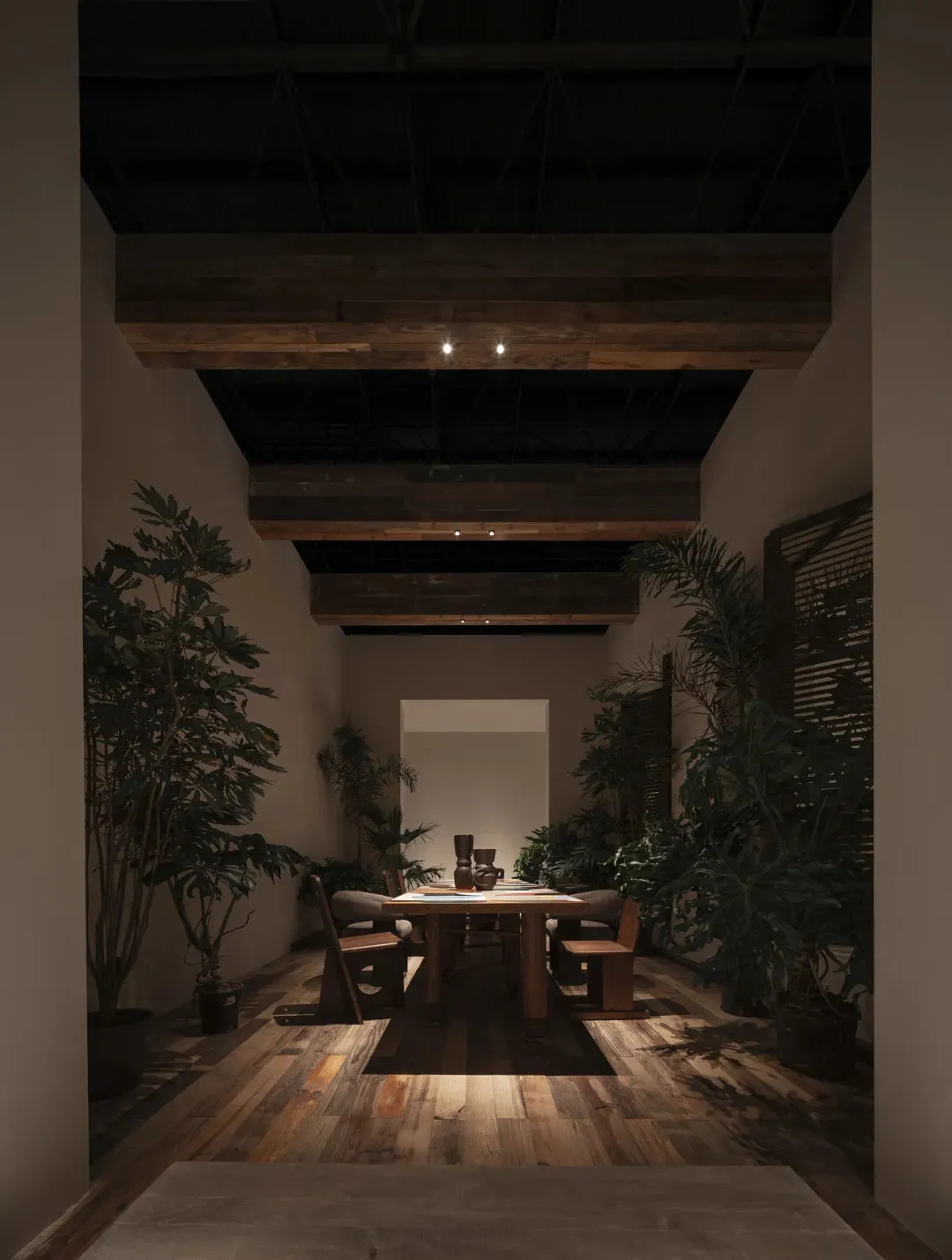 Long narrow dining room featuring massive reclaimed wood ceiling beams, a wooden dining table with matching chairs, and numerous large potted tropical plants under warm spotlighting.