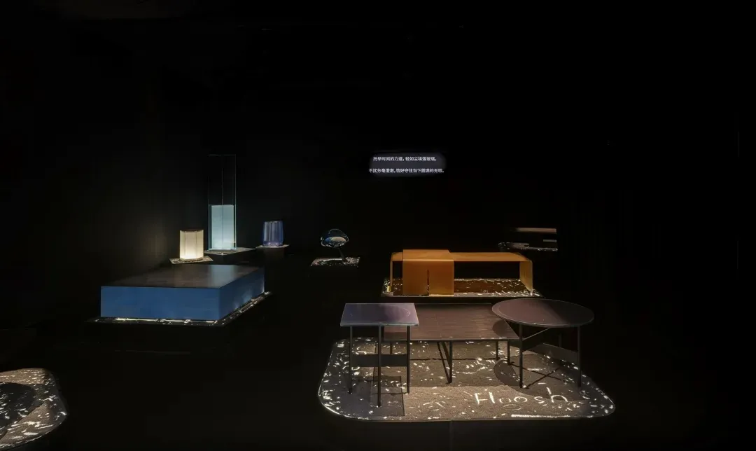 Dark exhibition space featuring modern furniture pieces on illuminated platforms, including a vibrant blue cube platform, an orange bench, and various tables under dramatic spotlighting.