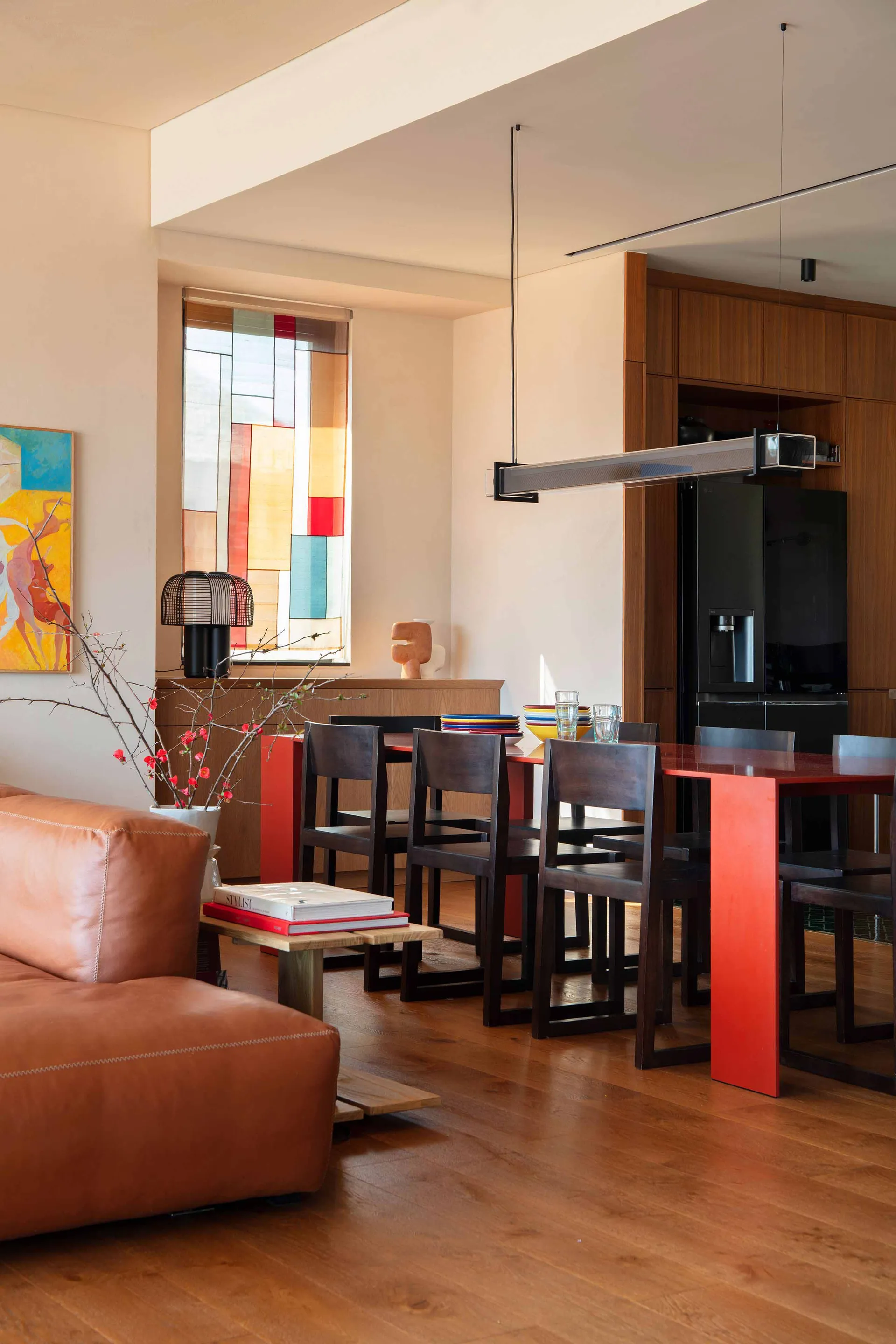 Modern dining area featuring a bright red dining table, dark walnut chairs, a brown leather sofa in the foreground, and colorful stained-glass window panels.