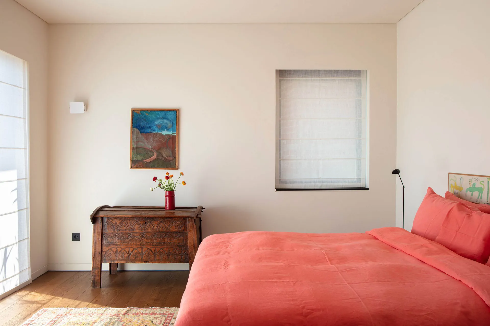 Bright bedroom with a coral-colored bed, antique carved wooden chest, colorful landscape painting, and natural light from two windows