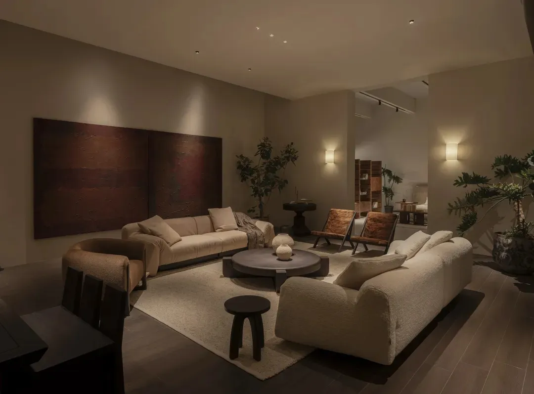 Modern living room featuring two boucle sofas, round coffee table, textured burgundy artwork, wooden accent chairs, and multiple indoor plants under warm ambient lighting.