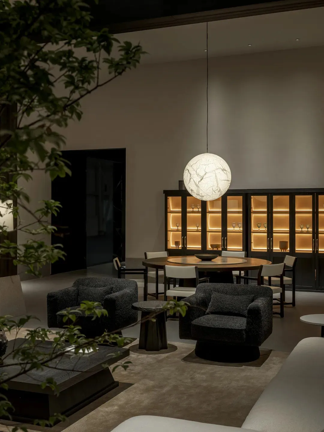 Modern luxury living and dining area featuring dark boucle swivel armchairs, a large spherical pendant lamp, round wooden dining table, and illuminated dark wood display cabinets.