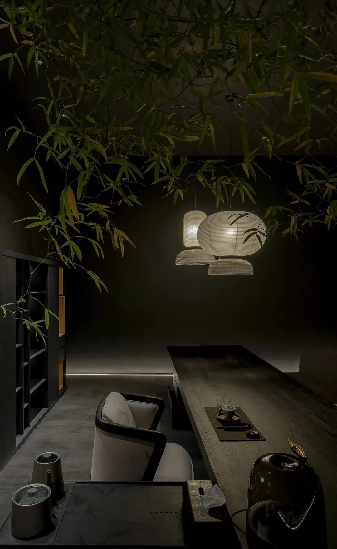 Modern Japanese-style tea room featuring a long dark wood table, light fabric armchairs, hanging paper lanterns, and fresh bamboo foliage hanging from the ceiling.