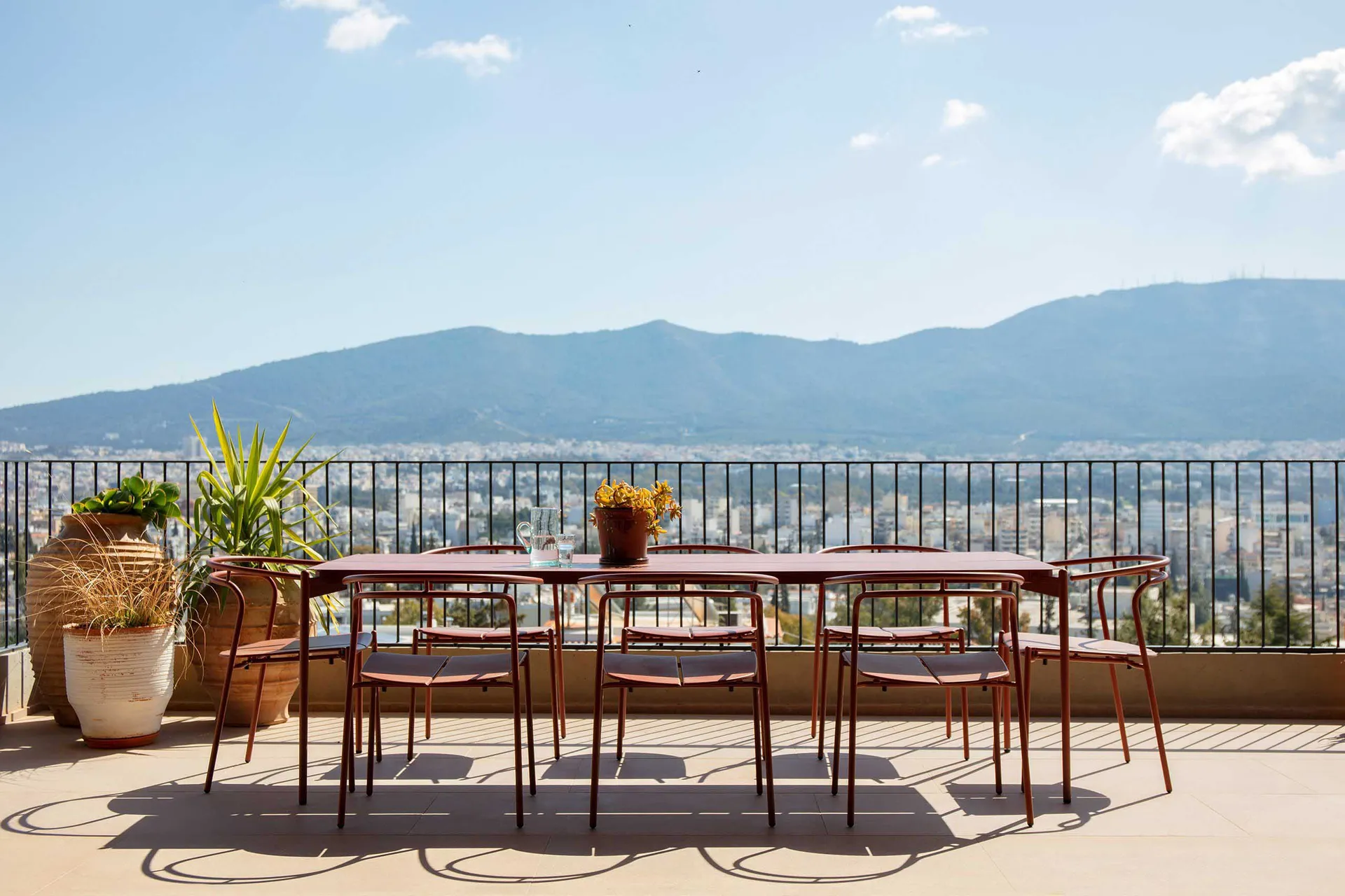 Outdoor dining table and six chairs in terracotta color on a rooftop terrace overlooking a vast mountain range and cityscape