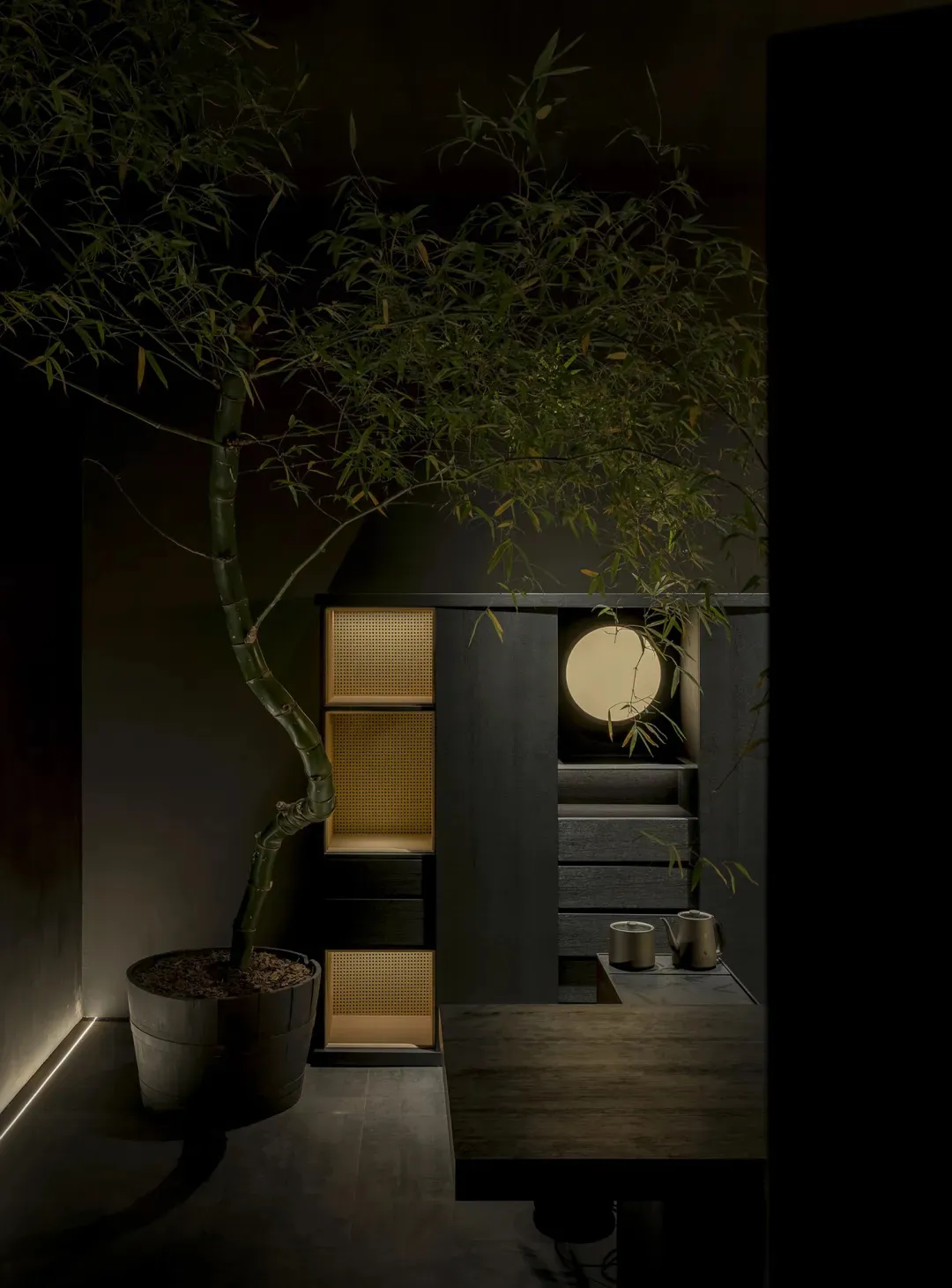 Modern minimalist tea room interior with a large potted bamboo tree, dark wood cabinetry with woven rattan panels, a glowing circular wall light, and a wooden table with tea set.