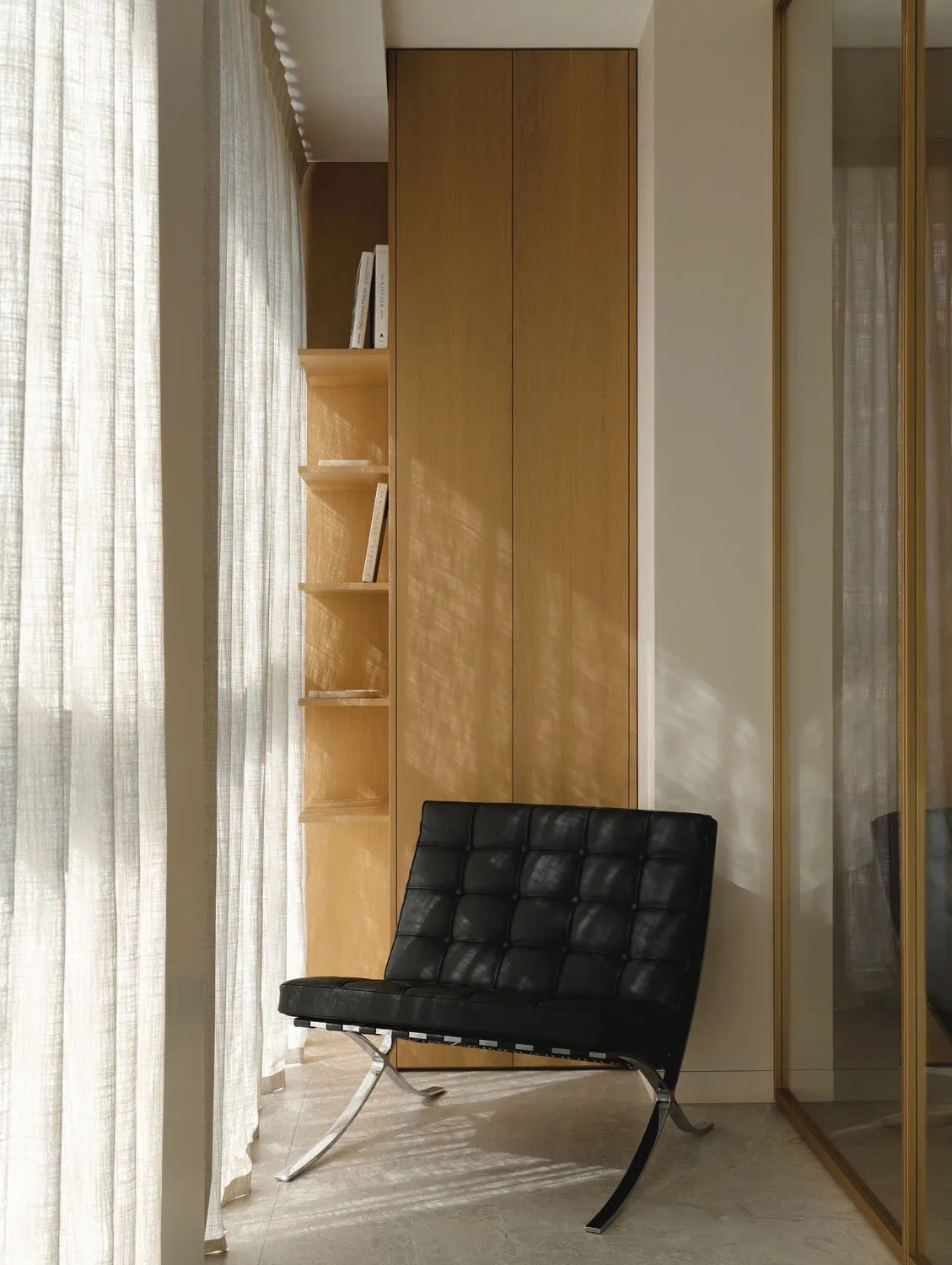 A black Barcelona chair placed in a bright minimalist reading nook with built-in oak shelving and sheer white curtains.