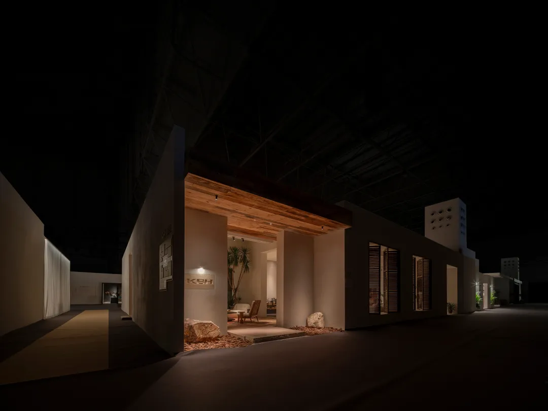 Night view of a modern exhibition booth inside a large exhibition hall, featuring a prominent reclaimed wood beam over the entrance, warm lighting, stone accents, and a glimpse of a contemporary living area with wooden furniture and plants.