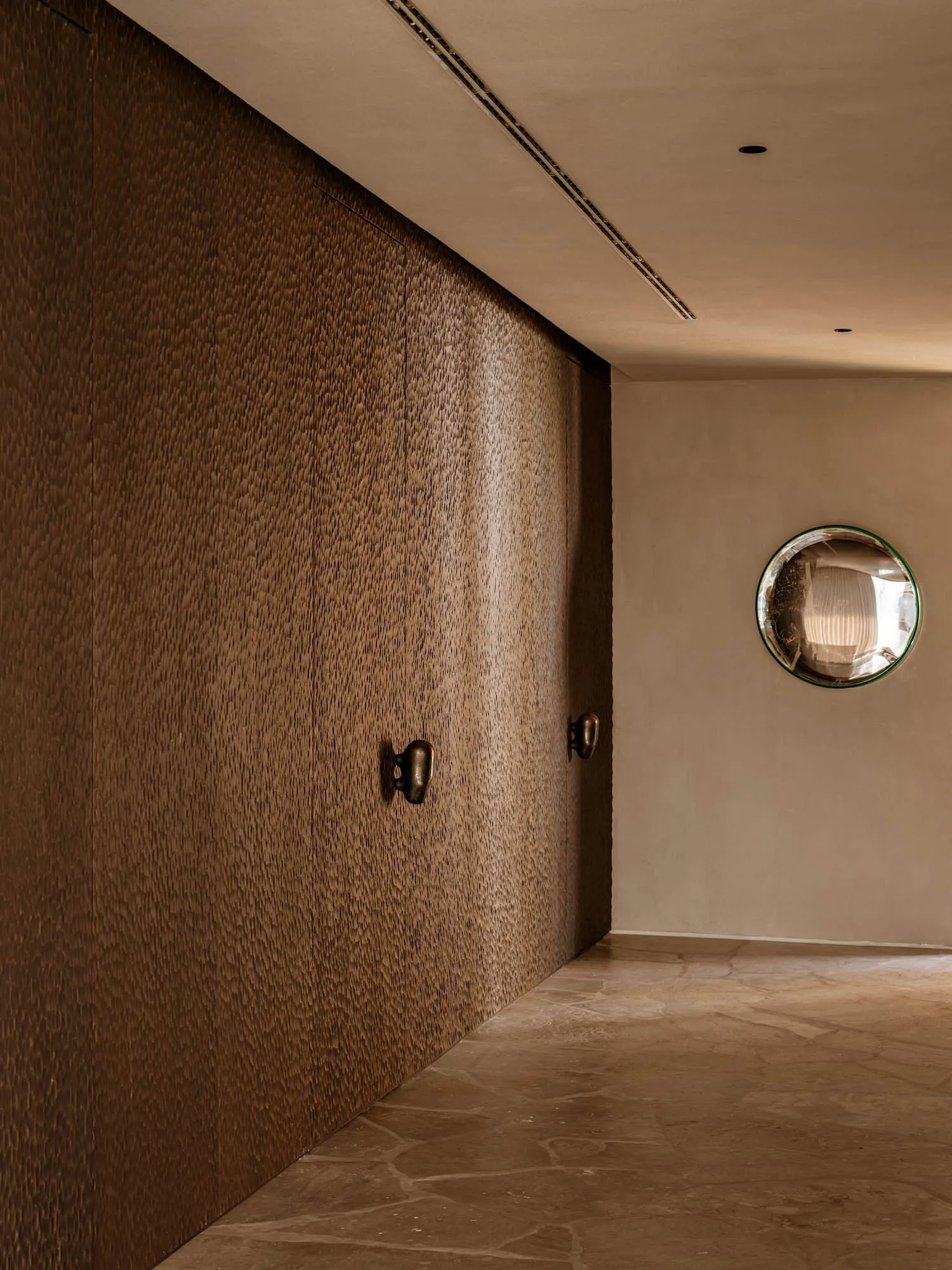 Modern minimalist hallway featuring a heavily textured brown wall, large-format travertine stone floor, and a round convex mirror on a neutral wall.