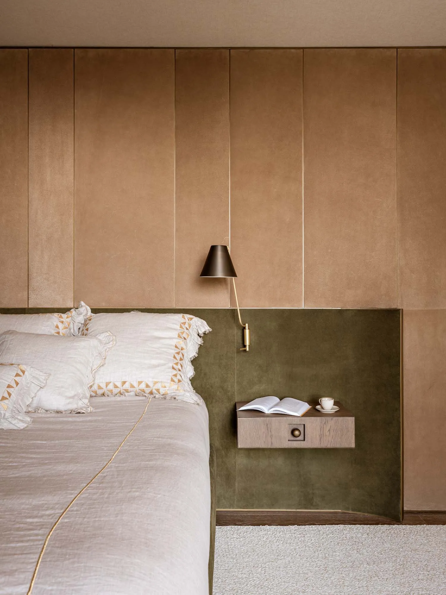 Modern minimalist bedroom featuring a warm beige upholstered wall, olive green velvet panel, floating wooden nightstand, brass wall lamp, and neutral linen bedding with geometric embroidery.