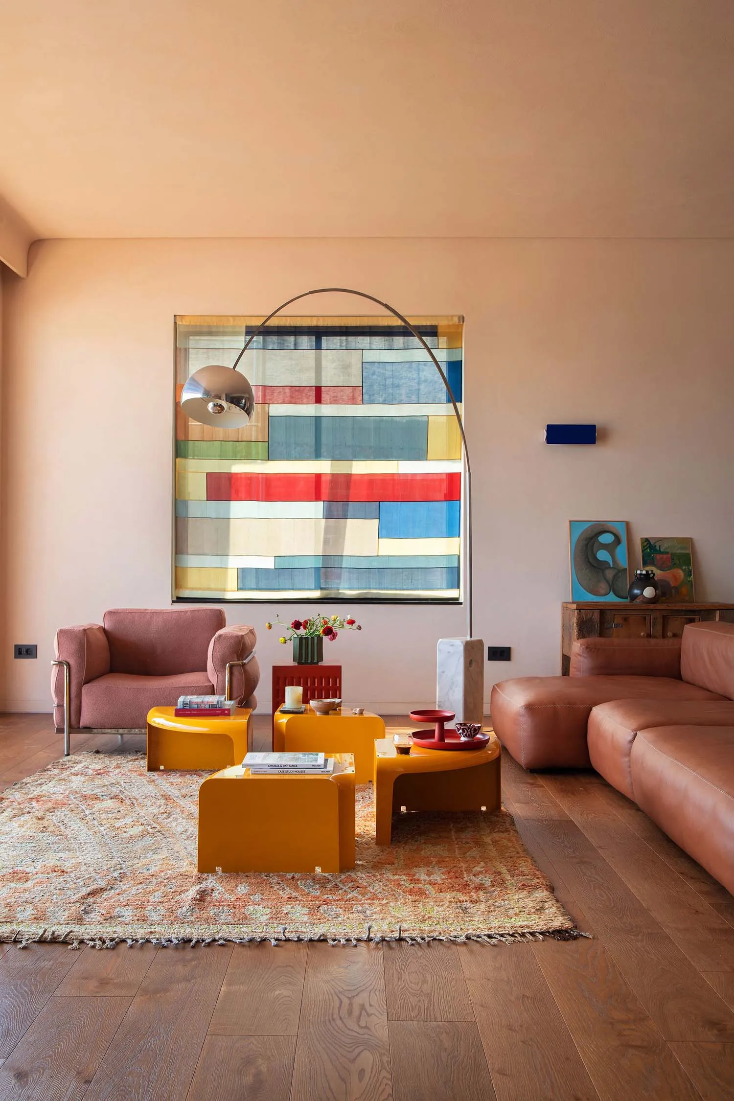 Bright living room with peach walls, a pink velvet armchair, brown leather sofa, glossy yellow nesting tables, a large colorful geometric tapestry, and a patterned area rug on wooden floors.