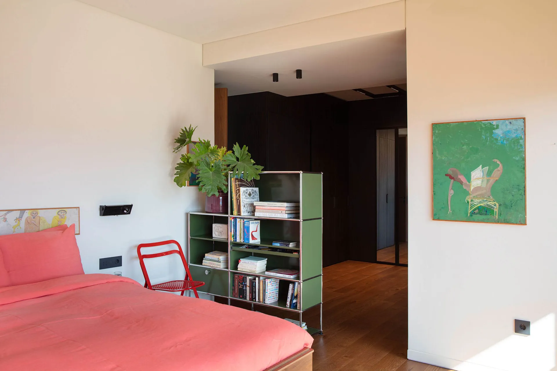 Modern bedroom featuring a coral pink bed, bright red metal chair, green modular bookshelf, and colorful abstract paintings