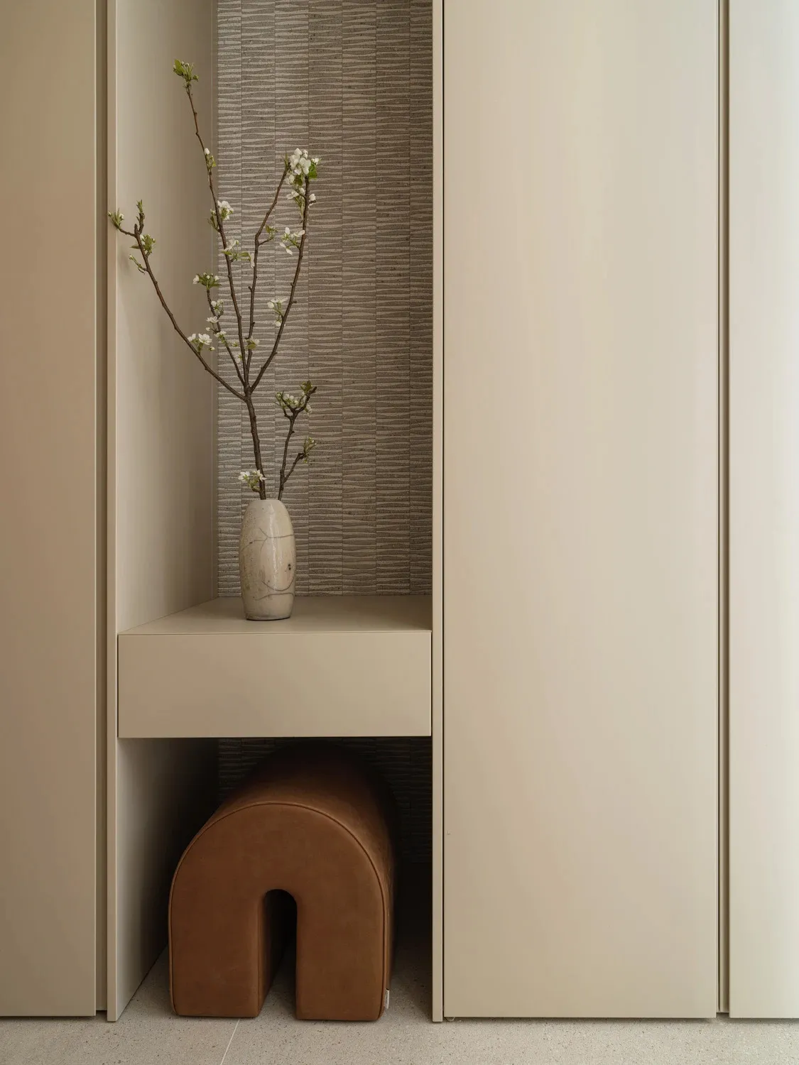 Modern minimalist entryway featuring a built-in beige niche with a crackle-glaze ceramic vase holding flowering branches, set against vertically textured neutral wallpaper, with a brown leather arched stool tucked underneath.