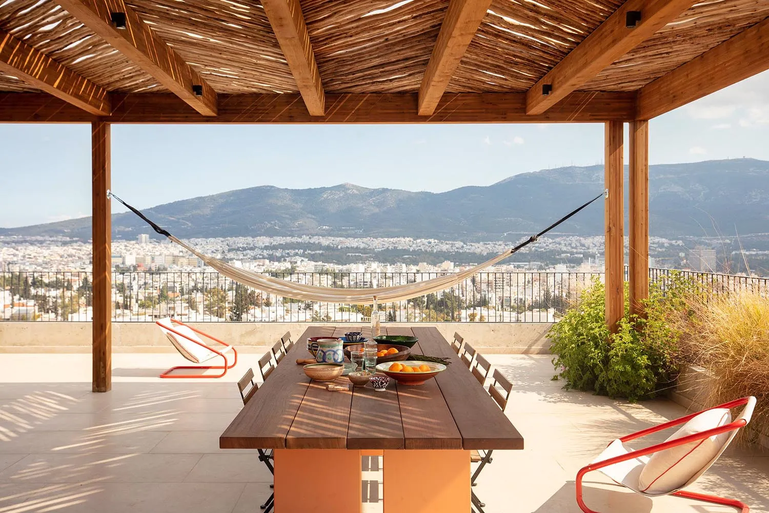 Outdoor rooftop terrace with a large teak wood dining table, red and white lounge chairs, a hanging hammock, and a panoramic view of mountains and a cityscape under a clear blue sky.
