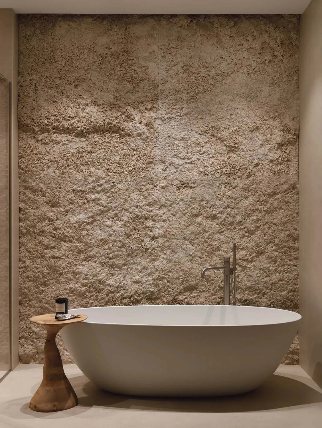Modern minimalist bathroom featuring a white freestanding bathtub, a textured travertine stone wall, and a natural wood side table with a candle.