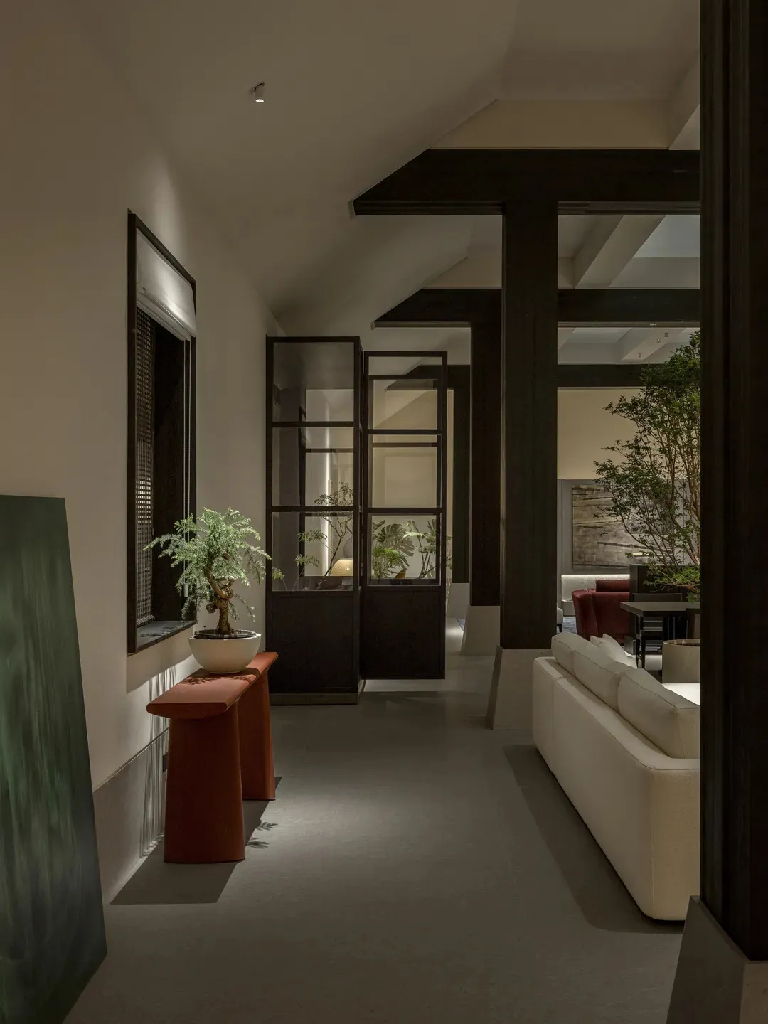 Modern minimalist hallway featuring dark wood structural beams, a white sectional sofa, terracotta console table with bonsai tree, and large green artwork.