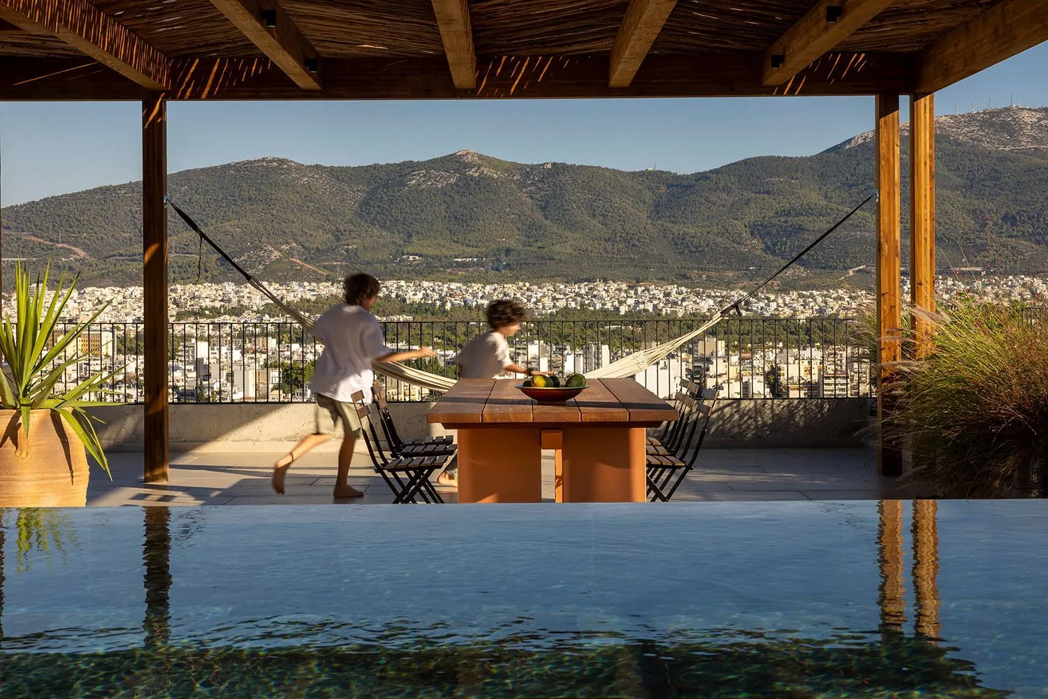 Two children playing on a wooden rooftop terrace beside an infinity pool, with a panoramic view of a city and mountains in the background under a clear blue sky.