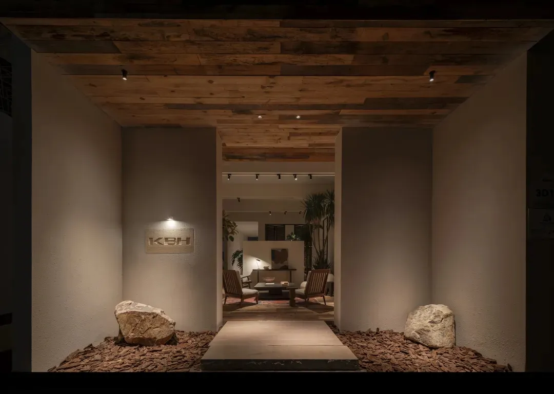 Modern entrance hallway featuring a reclaimed wood ceiling, textured beige walls, two large natural boulders on a bed of wood chips, and a view into a tropical-style living area beyond.