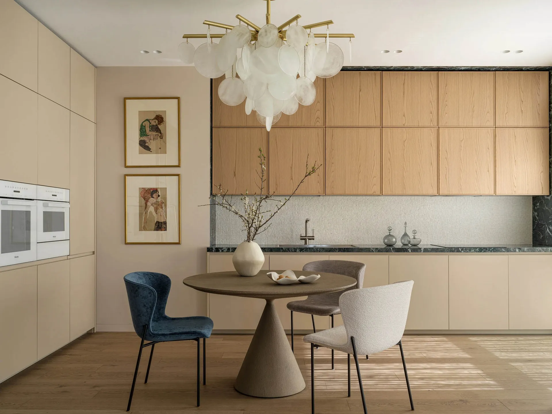 Modern dining area featuring a round concrete dining table, one blue velvet chair and two light textured fabric chairs, under a large white cloud-like chandelier, with light oak kitchen cabinetry in the background.