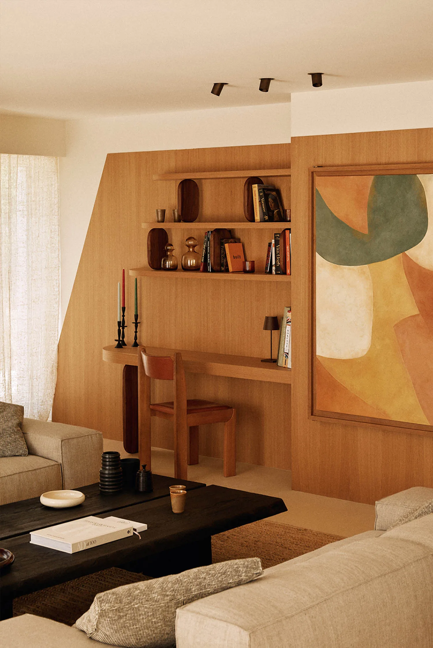 Modern living room featuring a built-in oak desk and shelving unit, large abstract painting, dark coffee table, and beige sofa with natural daylight.