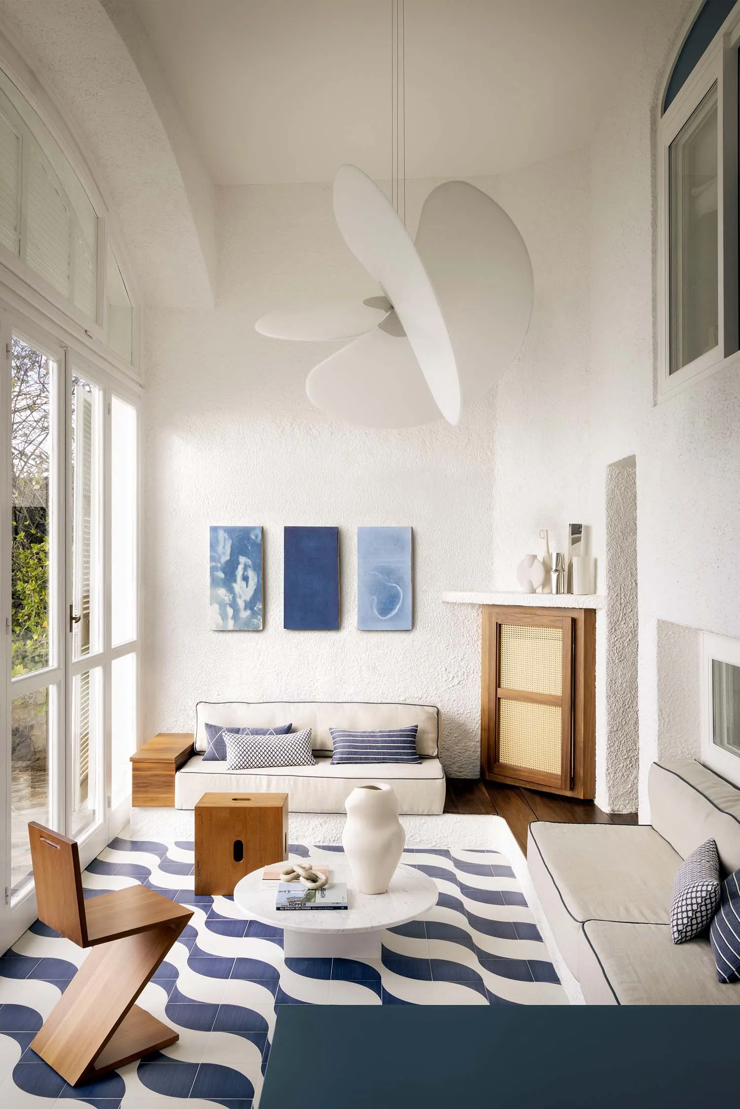 Bright sunroom interior with blue and white wave-pattern terrazzo floor tiles, white sofa, wooden zigzag chair, round marble coffee table, and large white sculptural pendant lamp.