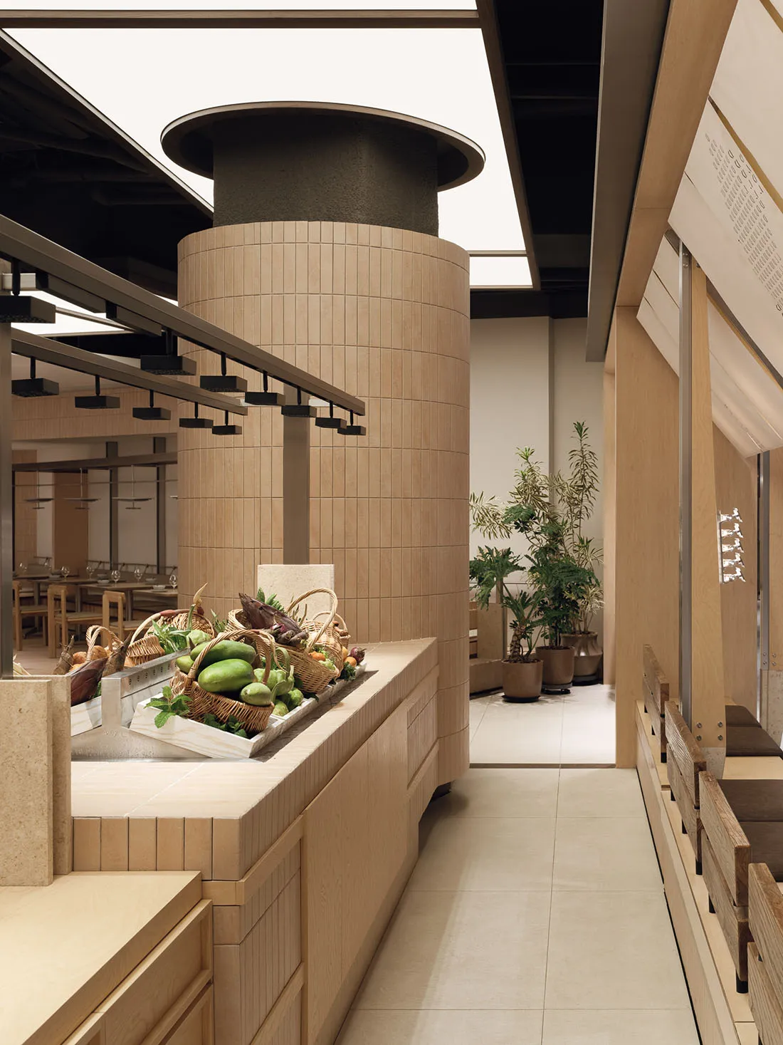Modern minimalist produce market interior with curved light-wood display counter filled with fresh fruits and vegetables in wicker baskets, large cylindrical concrete column with wood slat cladding, potted plants, and wooden bench seating along the right wall.