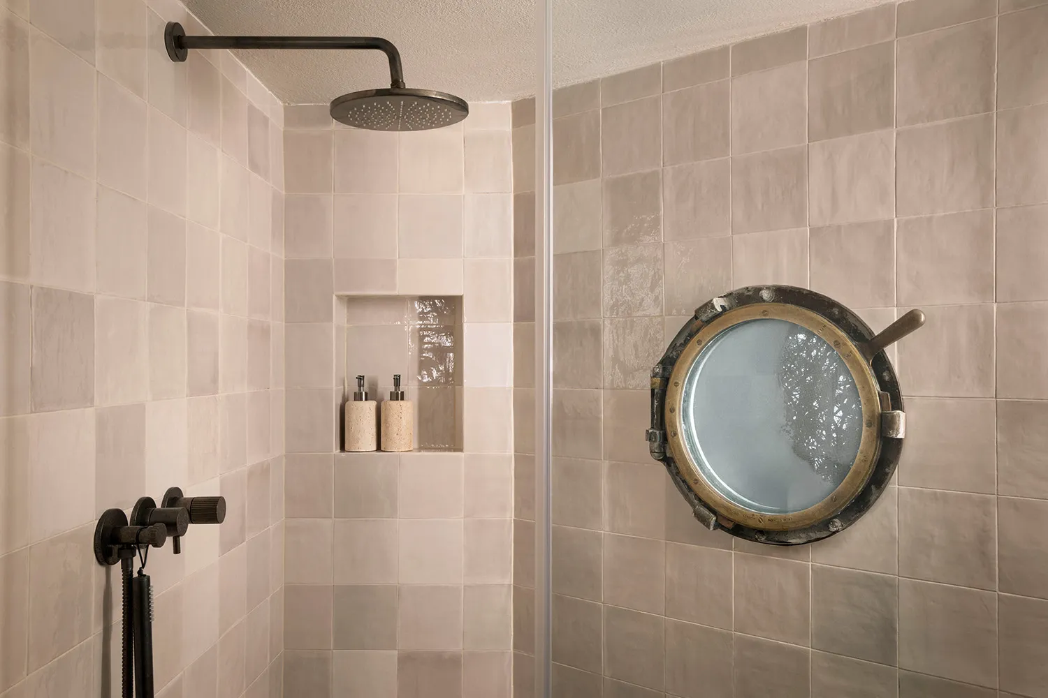Modern bathroom shower corner with large round brass porthole mirror, beige rectangular tiles, black rain shower head and matte black wall mixer.