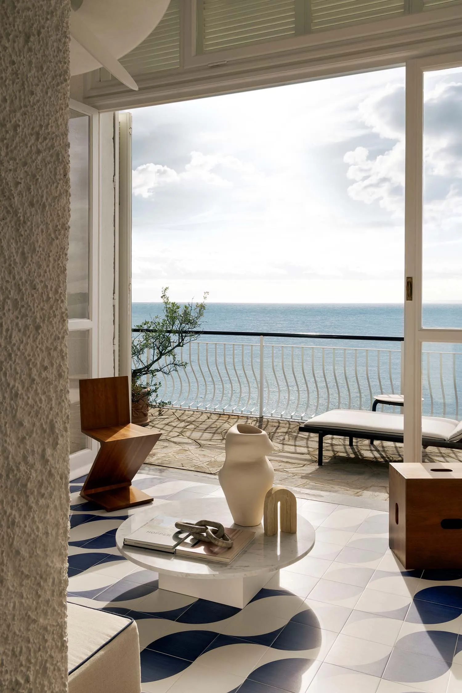 Modern coastal living space with blue and white patterned terrazzo floor tiles, white marble coffee table, wooden zigzag chair, and large open doors revealing a panoramic ocean view under a bright blue sky.