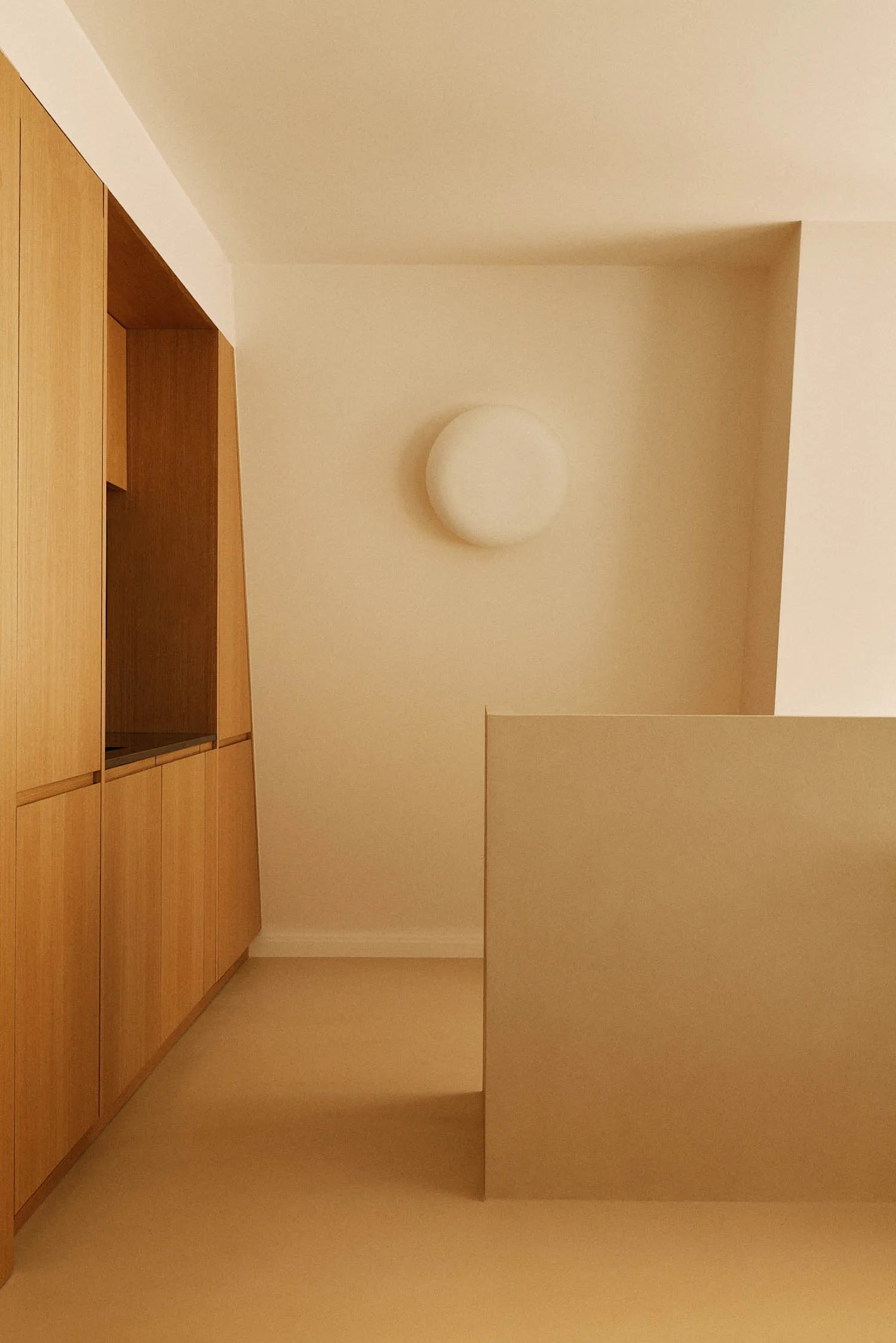 Minimalist entryway featuring light oak built-in cabinetry, hidden storage, a round white wall sconce, and a beige half-wall partition.