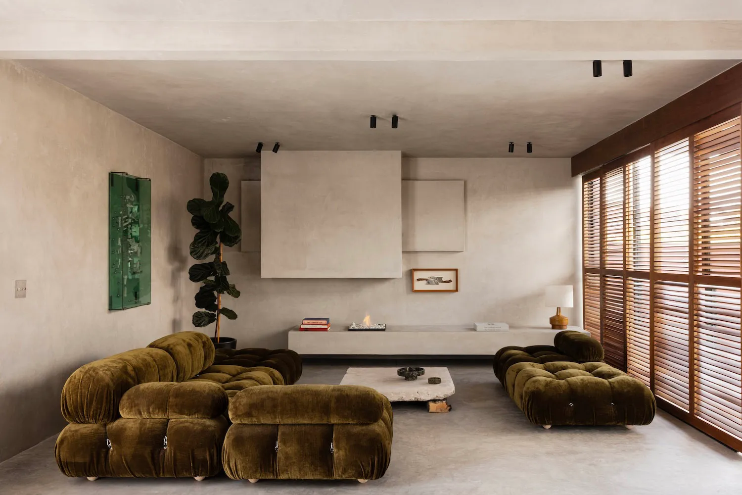 Modern minimalist living room featuring two olive green modular velvet sofas, a stone coffee table, concrete walls and ceiling, wooden window shutters, and a large potted fiddle leaf fig tree.
