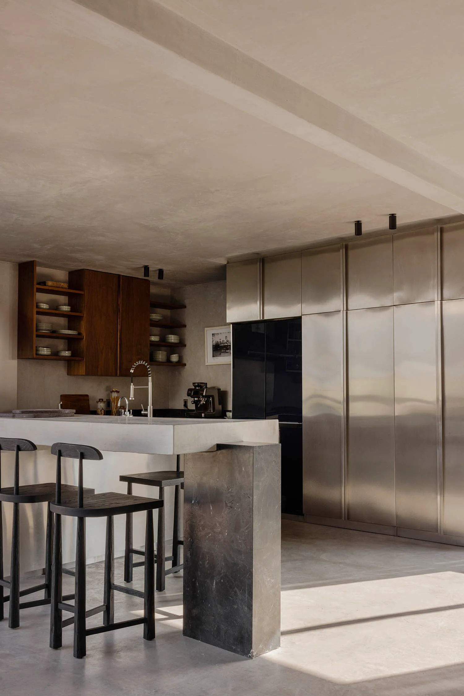 Modern minimalist kitchen with a heavy concrete island, three dark wooden bar stools, open walnut shelving, and large stainless steel cabinetry under a raw concrete ceiling.