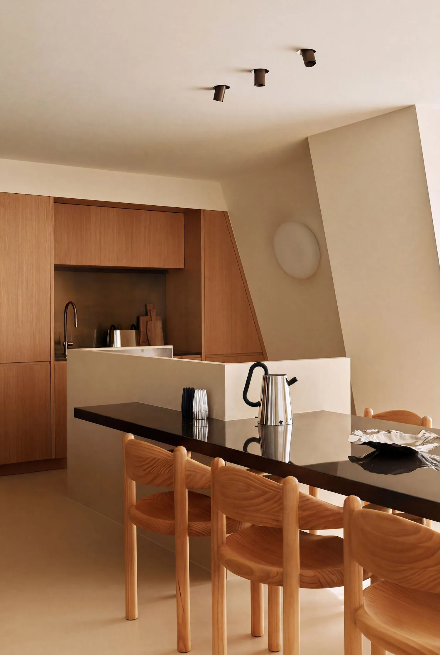 Modern minimalist dining area featuring light oak wooden chairs, a glossy black stone dining table, and a kitchen island with warm wood cabinetry in the background.