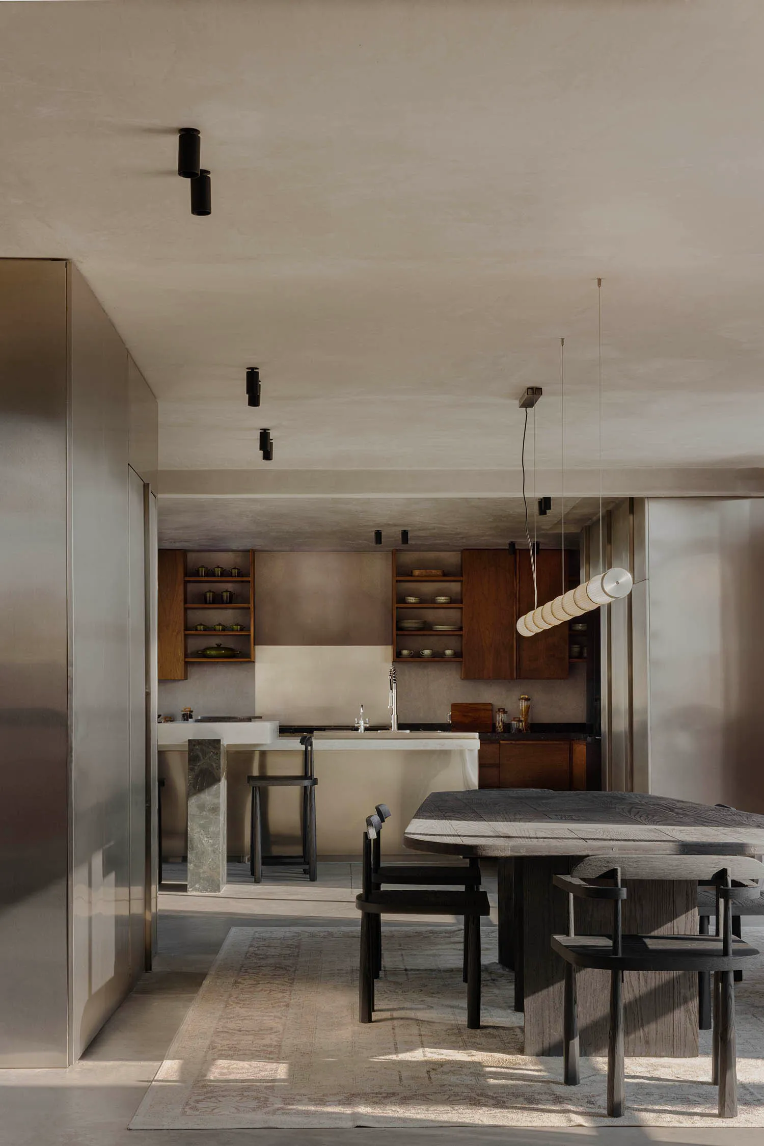 Modern minimalist open-plan kitchen and dining area featuring a dark walnut dining table, matching wooden chairs, concrete kitchen island, and warm walnut cabinetry under a sculptural cylindrical pendant light.