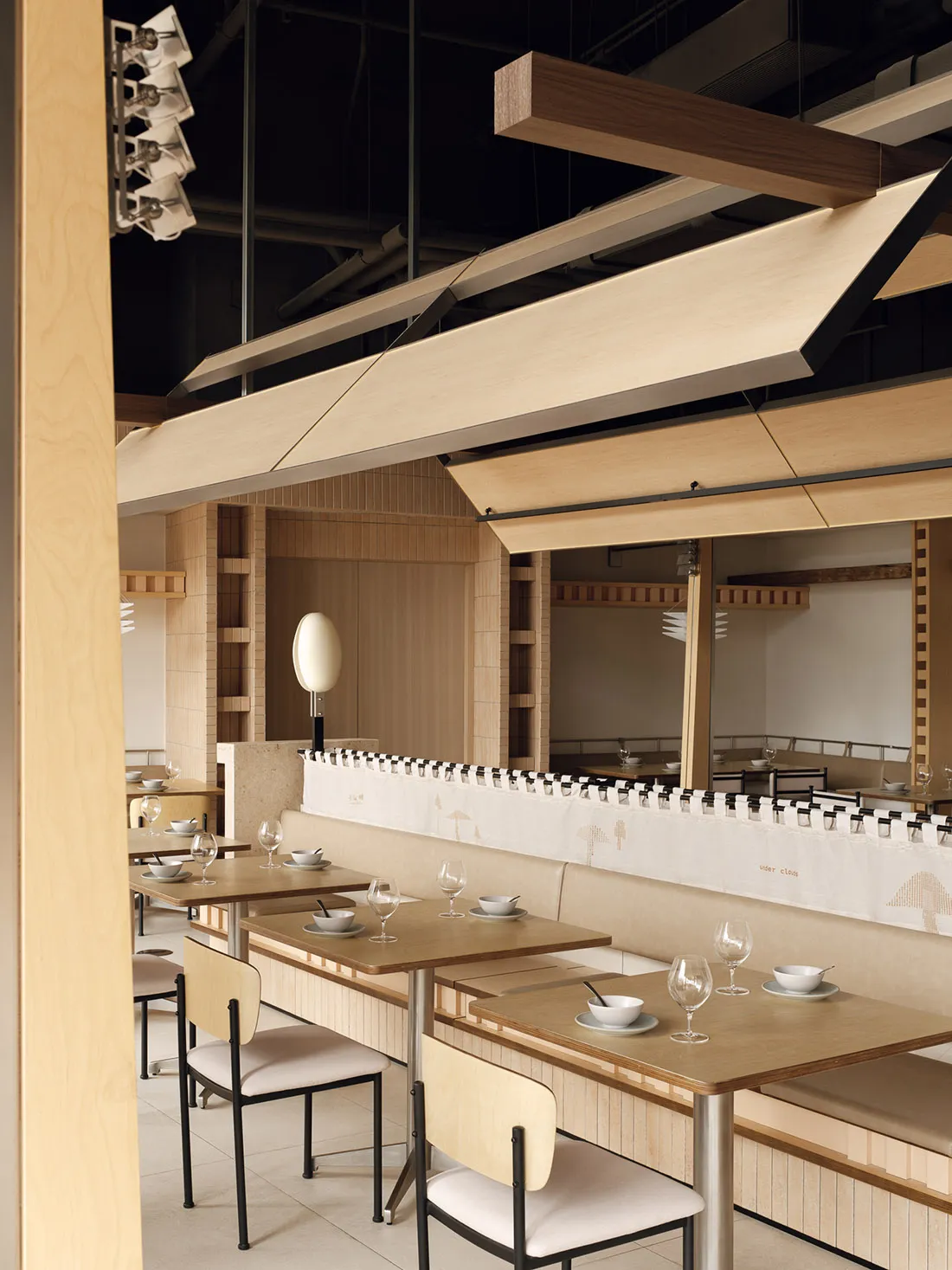 Modern minimalist restaurant interior featuring light oak wooden tables, upholstered booths, black metal chairs with bentwood backs, and a dramatic sculptural oak canopy hanging from the ceiling.