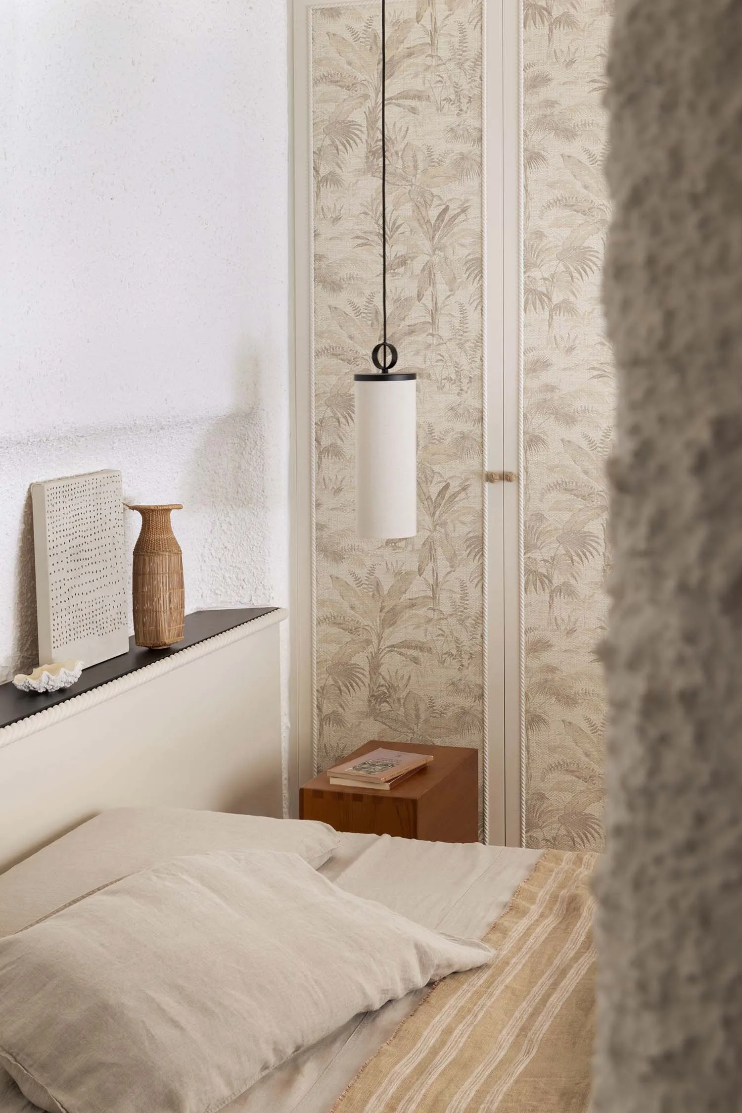 Cozy bedroom corner with beige linen bedding, wooden nightstand, cylindrical pendant lamp, and tropical leaf wallpaper in neutral tones.