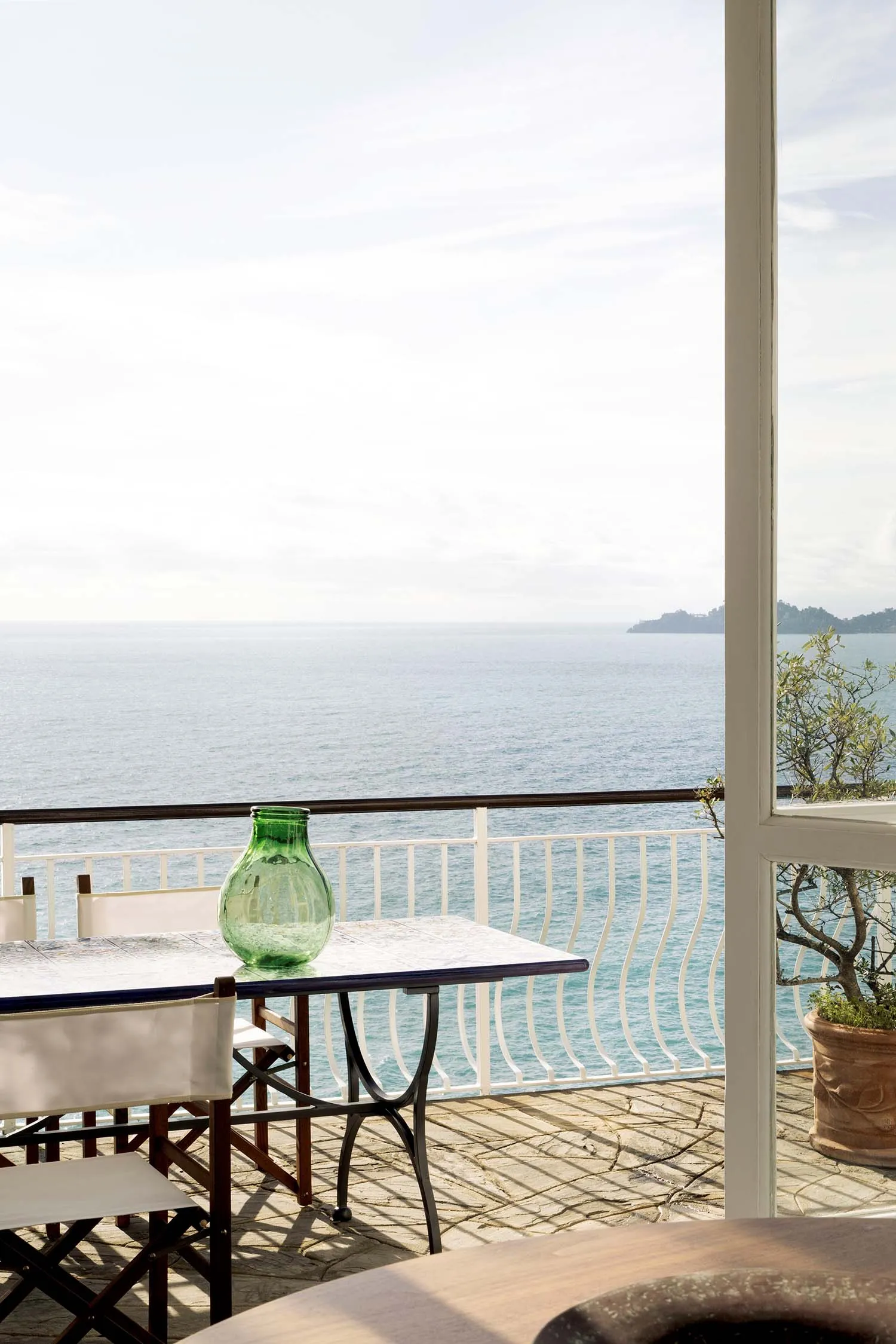 Outdoor stone balcony with white folding chairs, green glass vase, and sweeping ocean view under bright daylight