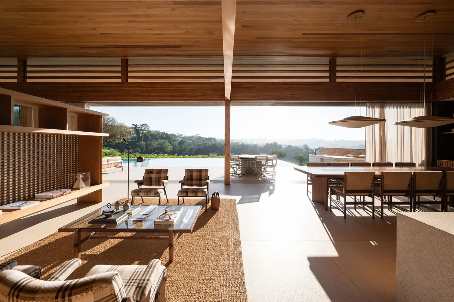 Bright open-plan living and dining area with warm wooden ceiling, plaid armchairs, large glass doors opening to a swimming pool and green hills.